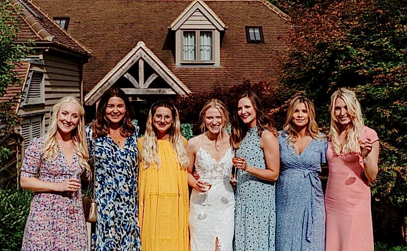 Hurley House - Seven women stand close together, smiling outdoors in front of a house. They wear colorful dresses, and one in the center is in white bridal attire, suggesting a wedding celebration. Sunlight and greenery surround them.