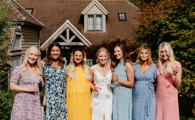 Hurley House - Seven women smiling in colorful dresses stand in front of a stunning luxury wedding venue in Berkshire, featuring a triangular roof and gabled windows. One woman, dressed in white, appears to be the bride. They are outdoors, surrounded by lush greenery and trees.