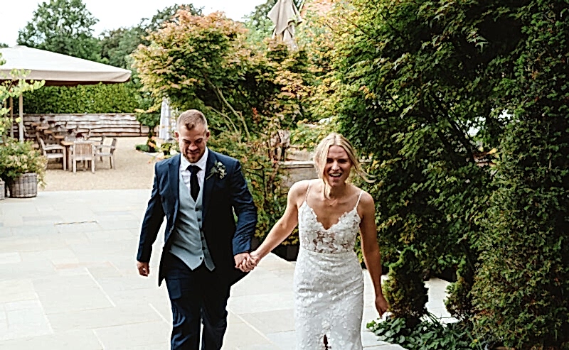 Hurley House - A joyful couple walks hand in hand on a stone path outdoors at a luxury wedding venue in Berkshire. The man is wearing a dark suit, and the woman is in a white wedding dress. They’re surrounded by lush greenery, and patio furniture is visible in the background.