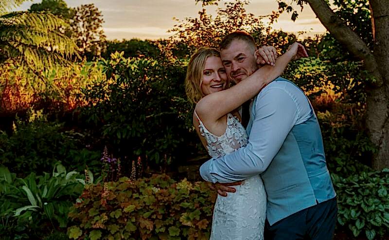 Hurley House - A couple embraces in a luxury wedding venue in Berkshires garden during sunset. The woman, in a white lace dress, smiles while the man, in a blue vest, hugs her tightly. They are surrounded by lush greenery and colorful plants, with a warm sky in the background.