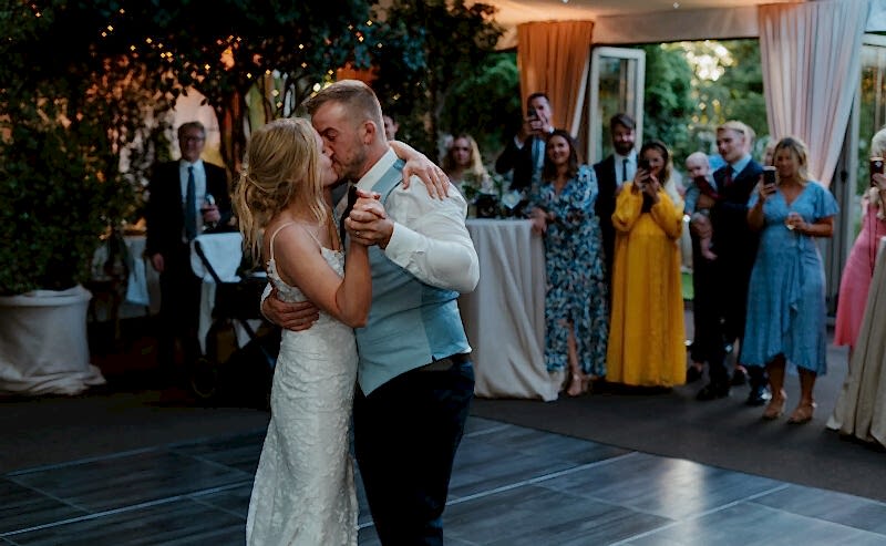 Hurley House - At a luxury wedding venue in Berkshire, a bride and groom share their first dance. Guests stand around watching, some taking photos. The elegantly decorated space, adorned with trees and draped curtains, creates a festive atmosphere perfect for this unforgettable moment.