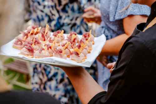 Hurley House - A person holds a white tray with neatly arranged appetizers topped with meat and garnish. People in patterned clothing are seen in the blurred background, suggesting a social or outdoor event.