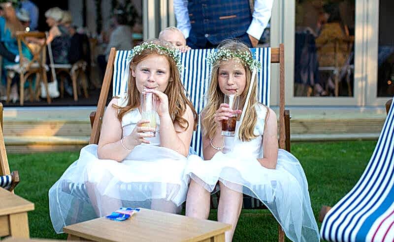 Hurley House - Two young girls in white dresses and floral headbands sit on striped deck chairs, sipping from bottles with straws. A baby peeks from the background, while a man in a vest stands nearby. This charming scene unfolds at a luxury wedding venue in Berkshire, surrounded by tables and guests outdoors.