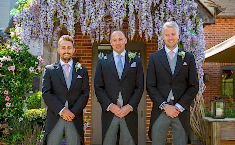 Hurley House - Three men in formal suits stand smiling in front of a brick building adorned with blooming wisteria, an exquisite backdrop for a luxury wedding venue in Berkshire. They each wear matching dark jackets, gray vests, and ties, complete with boutonnieres on their lapels.