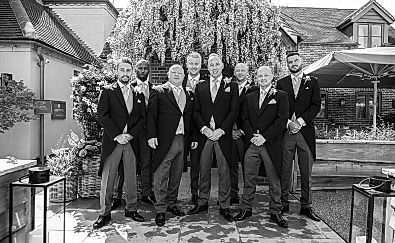 Hurley House - A group of eight men in formal suits stand together outdoors, posing for a photo at a luxury wedding venue in Berkshire. They are in front of a lush, white flowering tree on a sunny day, with brick buildings and a patio area elegantly framing the backdrop.
