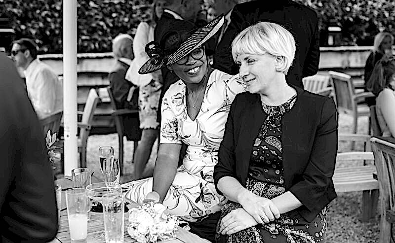 Hurley House - Two women sit and smile at each other outdoors during a social gathering at a luxury wedding venue in Berkshire. One wears a floral dress and hat, the other a patterned dress and blazer. Glasses of drinks are on the table before them, with people and foliage in the black-and-white background.