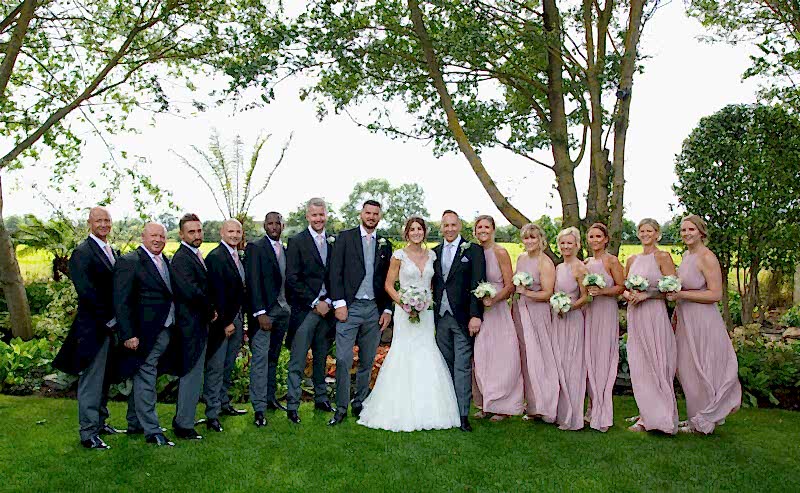 Hurley House - A wedding party poses outdoors at a luxury wedding venue in Berkshire, under lush trees. The bride, in a white gown, stands with the groom and groomsmen in gray suits on one side, while bridesmaids in lilac dresses are on the other. They all smile with greenery as their backdrop.