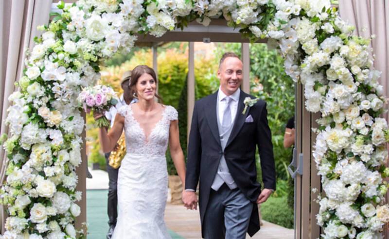 Hurley House - A couple strolls hand in hand under a floral archway adorned with white flowers at a luxury wedding venue in Berkshire, holding a bouquet of pink and purple blooms. The bride dazzles in a lace gown, while the groom looks sharp in a black suit with a white tie and boutonniere.