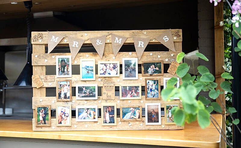 Hurley House - A wooden photo display board with Mr & Mrs at the top showcases a couples cherished moments, adorned with wooden hearts and burlap flags. Set on a kitchen counter with nearby plants, its a charming nod to their Luxury Wedding venue in Berkshire.