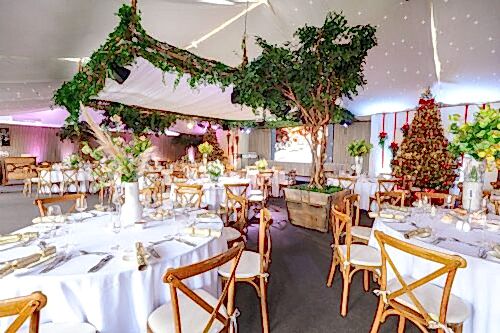 Hurley House - Elegant event space decorated with round tables set for dining, wooden chairs, lush green trees in planters, hanging greenery, and a Christmas tree with red ornaments, all under a white canopy with string lights.