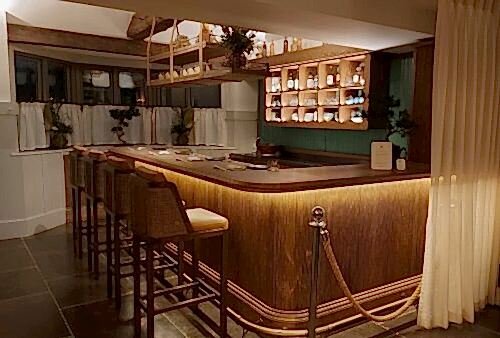 Hurley House - A cozy, dimly lit bar with a wooden counter and four high-backed chairs. Shelves with bottles and glasses are in the background, and hanging plants add a touch of greenery. A rope barrier and curtain frame the area.