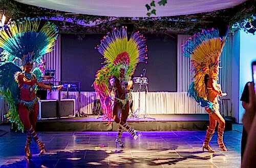 Hurley House - Three dancers in colorful feathered costumes perform energetically on a stage with vibrant lighting. The background has musical instruments and decorations with greenery above. An audience member is visible in the foreground.