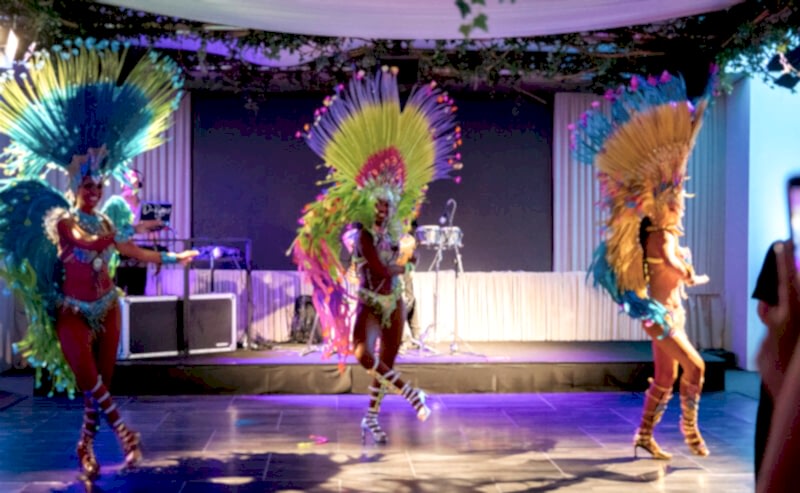 Hurley House - Three dancers in colorful feathered costumes perform energetically on a stage with vibrant lighting. The background has musical instruments and decorations with greenery above. An audience member is visible in the foreground.