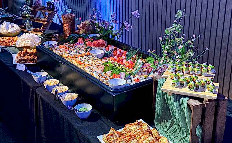 Hurley House - A buffet table displays a large variety of sushi rolls, sashimi, dumplings, and sauces, surrounded by decorative flowers and elegant tableware, set against a wooden wall with blue lighting.