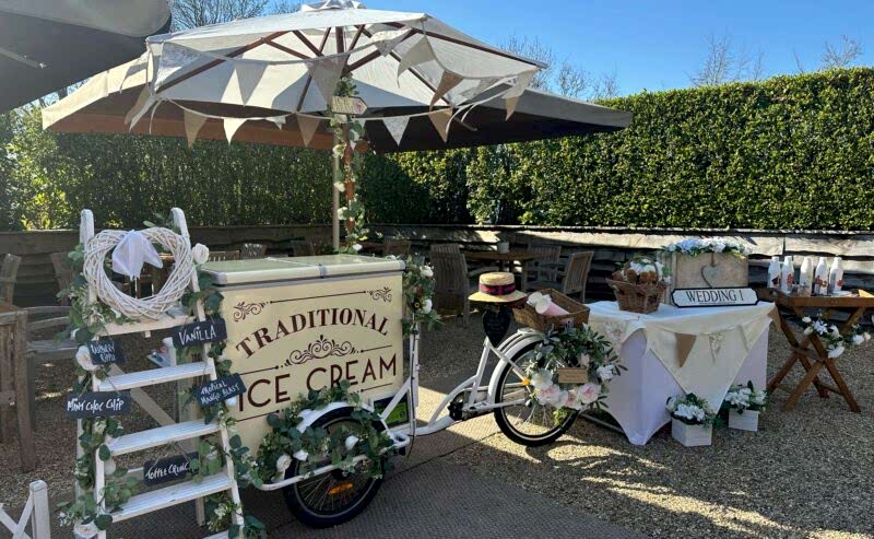 Hurley House - A vintage-style ice cream bike cart, featuring a Traditional Ice Cream sign, is set up outside under a large umbrella in a luxury wedding venue in Berkshire. Adorned with flowers and greenery, its accompanied by a wedding sign, small table, and decorated ladder in the sunny garden setting.