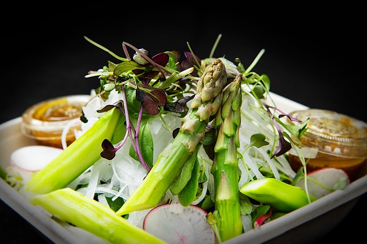 Hurley House - A close-up of a fresh salad featuring asparagus spears, radish slices, microgreens, and shredded vegetables, served in a container with two small cups of dressing in the background.