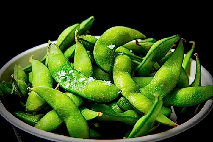 Hurley House - A close-up of a bowl filled with fresh green edamame pods, lightly sprinkled with coarse sea salt, against a dark background.