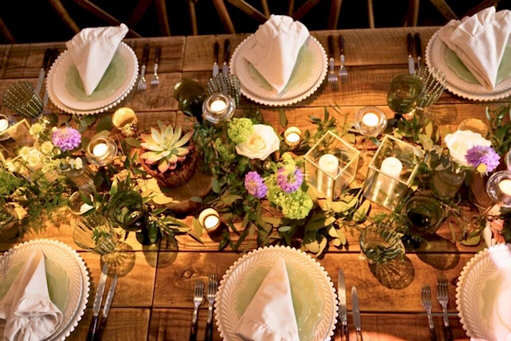 Hurley House - A rustic wooden table at this luxury wedding venue in Berkshire is elegantly set with white plates, neatly folded napkins, and green-tinted glasses. The centerpiece features candles, succulents, and a mix of purple and green flowers, creating a warm, inviting atmosphere.