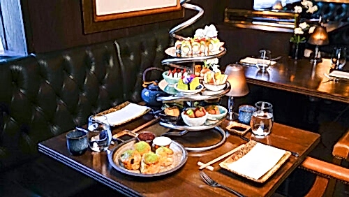Hurley House - A stylish restaurant table set for two, featuring an elaborate metal stand with tiers of assorted sushi, small dishes, and desserts, along with teapots, cups, and plates arranged neatly on a dark wooden table.