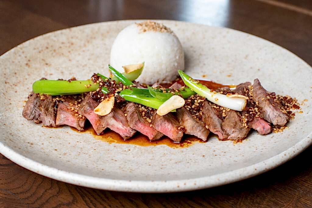 Hurley House - A plate with sliced seared steak topped with green onions, garlic, and sauce, served with a round mound of white rice on a speckled ceramic dish, set on a wooden table.
