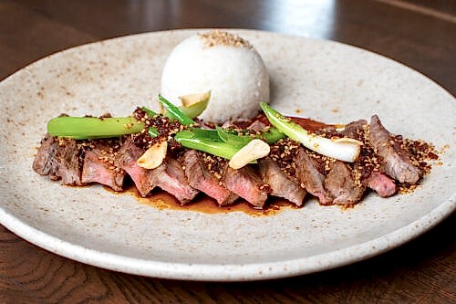Hurley House - A plate with sliced seared steak topped with green onions, garlic, and sauce, served with a round mound of white rice on a speckled ceramic dish, set on a wooden table.