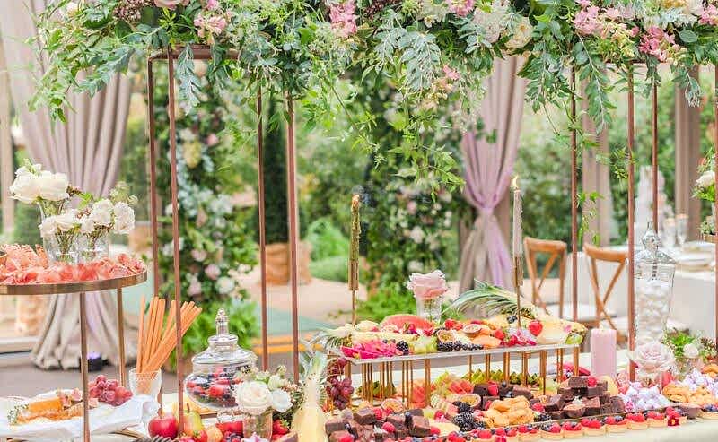 Hurley House - A beautifully arranged charcuterie board graces the wedding buffet at a luxury venue in Berkshire, filled with fruits, cheeses, and meats. Elegant floral decorations overhead feature pink and white roses with greenery, set against the backdrop of soft drapes and a picturesque garden.