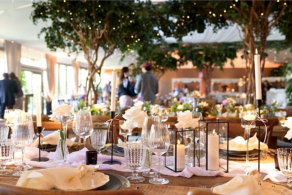 Hurley House - Elegant wedding table setting at a luxury wedding venue in Berkshire, featuring crystal glasses, white rose centerpieces, and lit candles in glass holders. Soft lighting and blurred guests in the background create a warm, festive atmosphere under lush greenery and fairy lights.