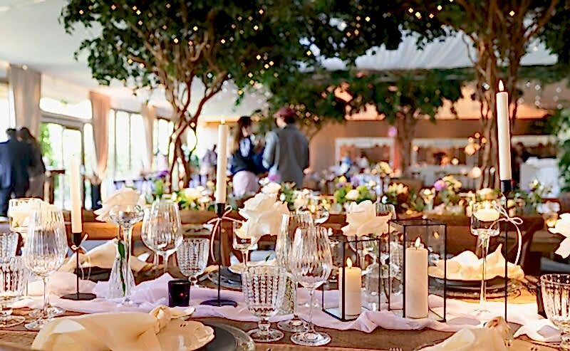 Hurley House - Elegant wedding table setting at a luxury wedding venue in Berkshire, featuring crystal glasses, white rose centerpieces, and lit candles in glass holders. Soft lighting and blurred guests in the background create a warm, festive atmosphere under lush greenery and fairy lights.