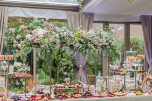 Hurley House - A lavishly decorated buffet table with an abundant spread of fruits, cheeses, sweets, and snacks. The table is topped with an elegant arrangement of fresh pink and white flowers and greenery, set in a bright, airy indoor setting.